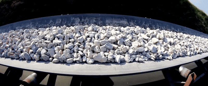 Weiße, gebrochene Steine werden auf einem Förderband transportiert. Die Steine sind gleichmäßig verteilt, das Förderband läuft durch eine industrielle Umgebung im Freien.